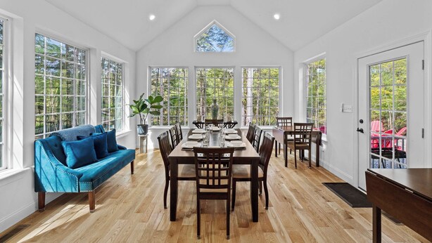 A dining area with a wooden table set for six, surrounded by chairs, a blue sofa, and large windows offering a view of trees. - Lakefront luxury,GameRoom,FirePit,Canoe,10Min2JFBB (Blakeslee)