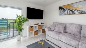 A modern living room with a grey sofa, a flat-screen TV mounted on the wall, a potted plant, and a large window offering a view of the outdoors. - Downtown OC Condo with Bay View, Pool, Parking, Walk to Beach & Boardwalk (Ocean City)