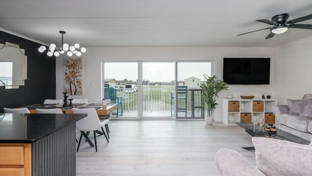 A modern living room with a large sliding glass door leading to a balcony, a kitchen area with a black countertop, and a grey sofa. - Downtown OC Condo with Bay View, Pool, Parking, Walk to Beach & Boardwalk (Ocean City)