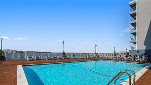 A rectangular swimming pool with a wooden deck, surrounded by lounge chairs and a white fence. - Downtown OC Condo with Bay View, Pool, Parking, Walk to Beach & Boardwalk (Ocean City)