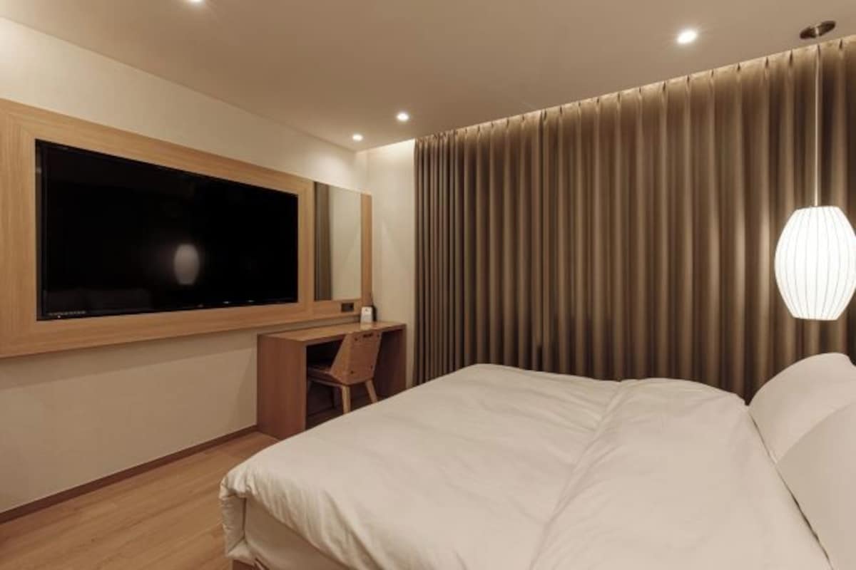 Habitación de hotel con una cama grande, un escritorio con silla, un televisor de pantalla plana y un amplio ventanal con cortinas.