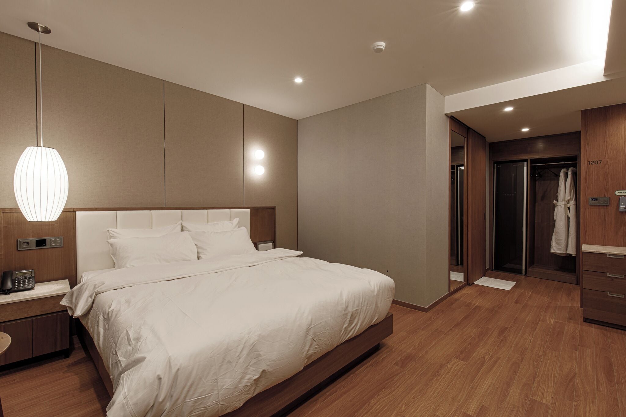 Une chambre d’hôtel moderne avec un grand lit, un téléphone sur la table de chevet, une petite table de nuit, un luminaire suspendu et une armoire en bois.
