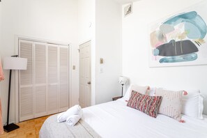 A bedroom with a bed, white pillows, a rolled towel, a nightstand, a lamp, and a door with a closet.