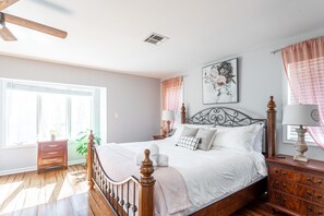 A bedroom with a wooden bed, a nightstand, a dresser, a ceiling fan, a window with curtains, and a wall-mounted lamp.