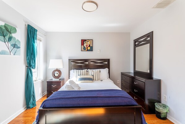 A bedroom with a bed, a bedside table with a lamp, a dresser, a mirror, and a window with curtains.