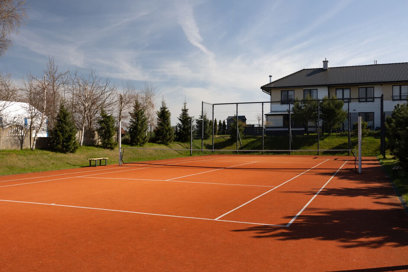 Una cancha de tenis con una red, rodeada de árboles y un edificio al fondo.