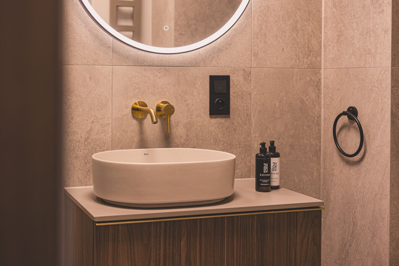 Une salle de bain avec un miroir rond, un lavabo, deux flacons de produits de toilette et un porte-serviette.