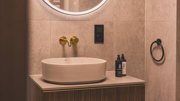 Une salle de bain avec un miroir rond, un lavabo, deux flacons de produits de toilette et un porte-serviette.