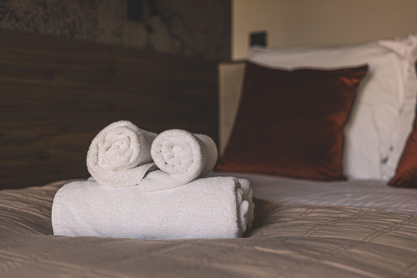 Una cama con toallas blancas enrolladas y una almohada marrón.
