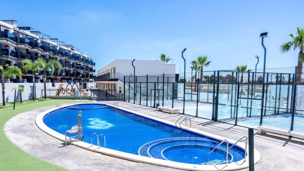 A swimming pool with a surrounding deck, a glass fence, and a building with balconies in the background.