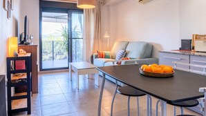 A modern living room with a sofa, a dining table with a fruit basket, a TV, and a large window with curtains.