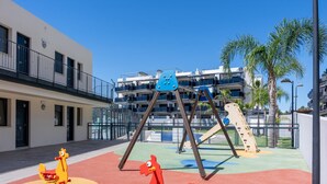 A playground with swings and a slide, surrounded by a building with balconies and palm trees.