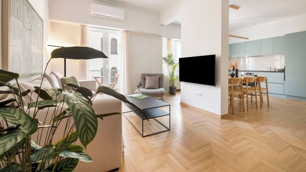 A modern living room with a sofa, coffee table, and TV. There is a dining area with chairs and a kitchen in the background. - Chic & Spacious 3BR Apartment at National Museum (Athina)
