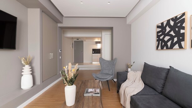 A modern living room with a grey sofa, a wooden coffee table, and a grey armchair. There is a flat-screen TV mounted on the wall and a decorative vase with dried plants. - Monastiraki Living: Deluxe 2BR Apartment (Athens)