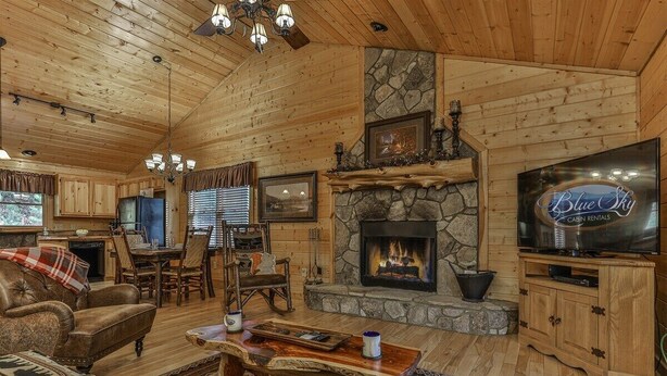 A log cabin interior with a stone fireplace, wooden furniture, a television, and a dining area.
