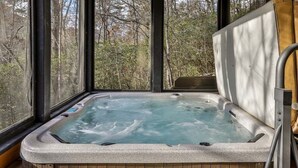 A hot tub with a wooden deck and a view of trees through large windows.