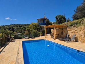 A clear blue pool with lounge chairs and a wooden pergola, surrounded by stone walls and greenery.