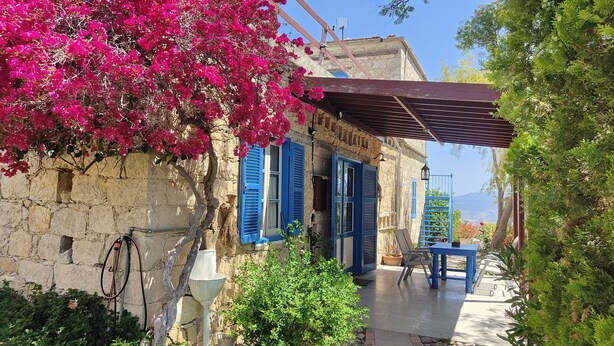 A rustic stone building with blue shutters, a covered outdoor seating area, and a garden with bougainvillea.