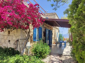Un edificio rústico de piedra con contraventanas azules, una zona de estar cubierta al aire libre y un jardín con buganvillas.