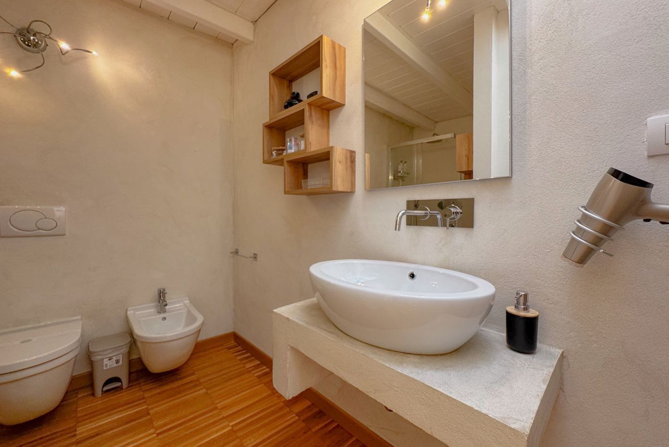 Un baño con un lavamanos grande y blanco, un bidé, un inodoro, estantes de madera, un espejo y un dispensador de toallas.