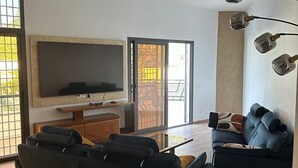 Ein modernes Wohnzimmer mit einem großen Flachbildfernseher, einem schwarzen Ecksofa, einem blauen Sessel und einem Glastisch.