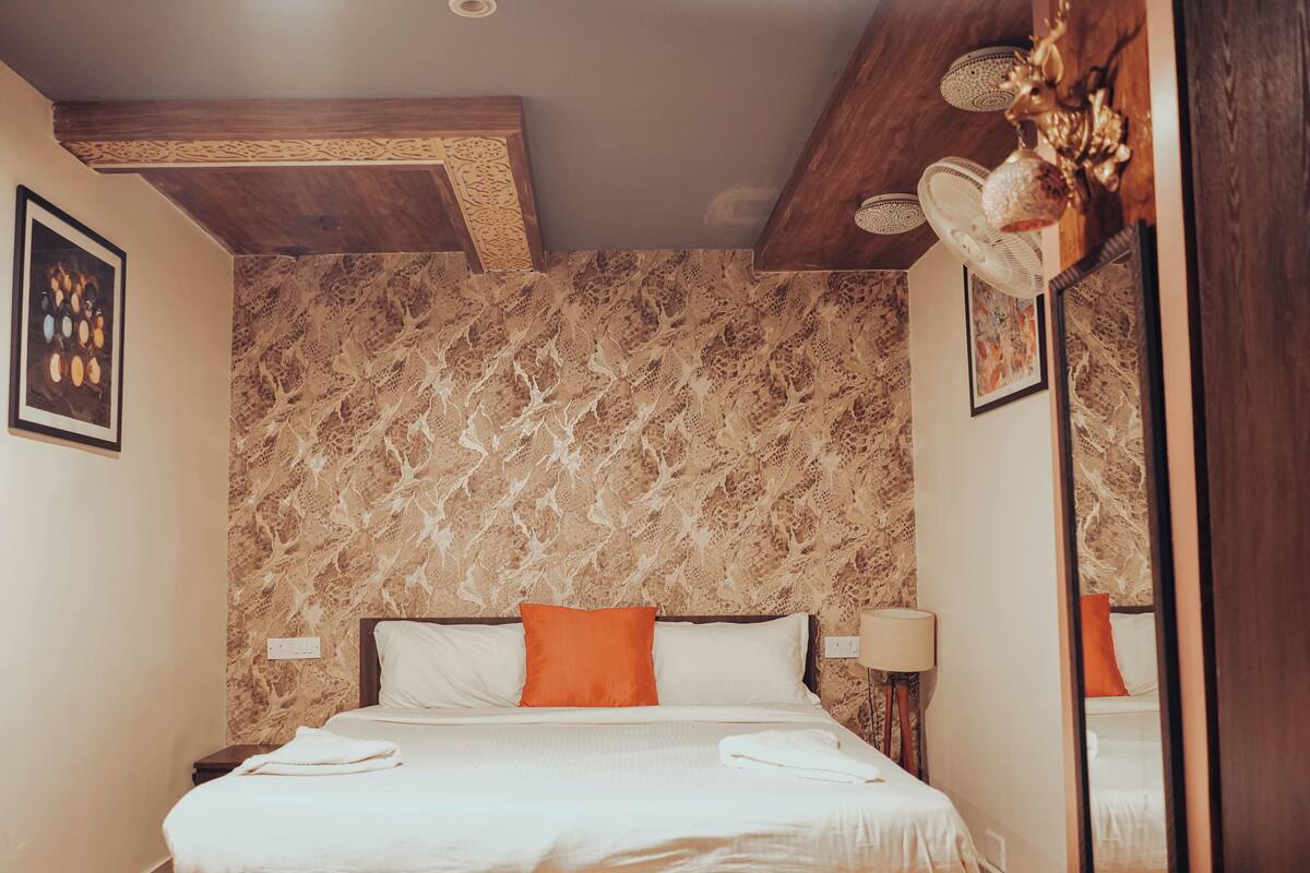 A hotel room with a marble-patterned wall, a wooden ceiling, a bed with white bedding and orange pillows, a bedside lamp, and a framed picture on the wall.