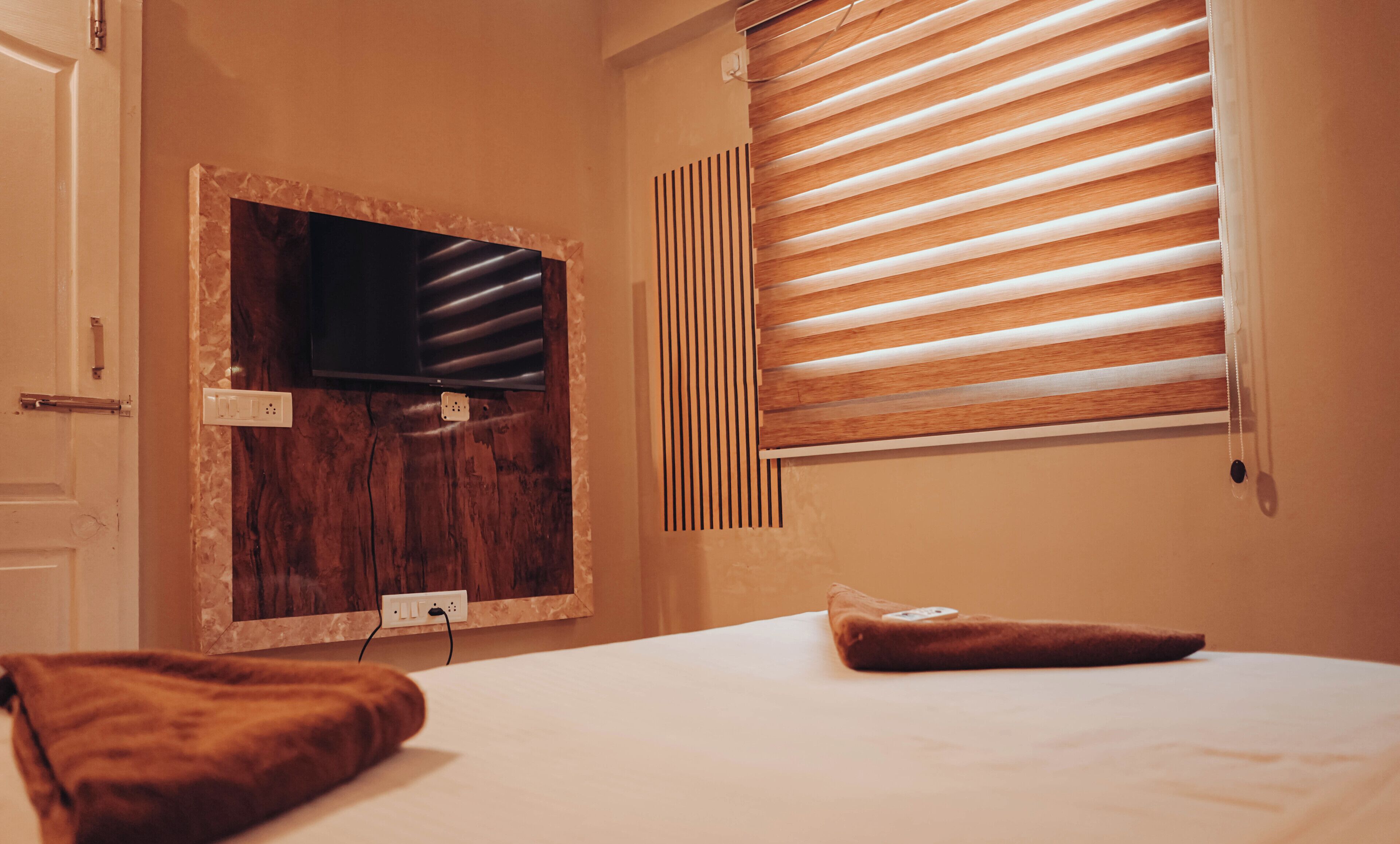 A bedroom with a wooden headboard, a window with blinds, and a television mounted on the wall.