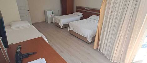 A hotel room with two single beds, a wooden desk with a telephone and towels, a wardrobe, a small refrigerator, and a window with curtains.