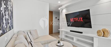 A modern living room with a large flat-screen TV, a beige sofa, a coffee table, and a wall-mounted shelf.