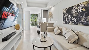 A modern living room with a large flat-screen TV, a sofa, a coffee table, and a dining area in the background.