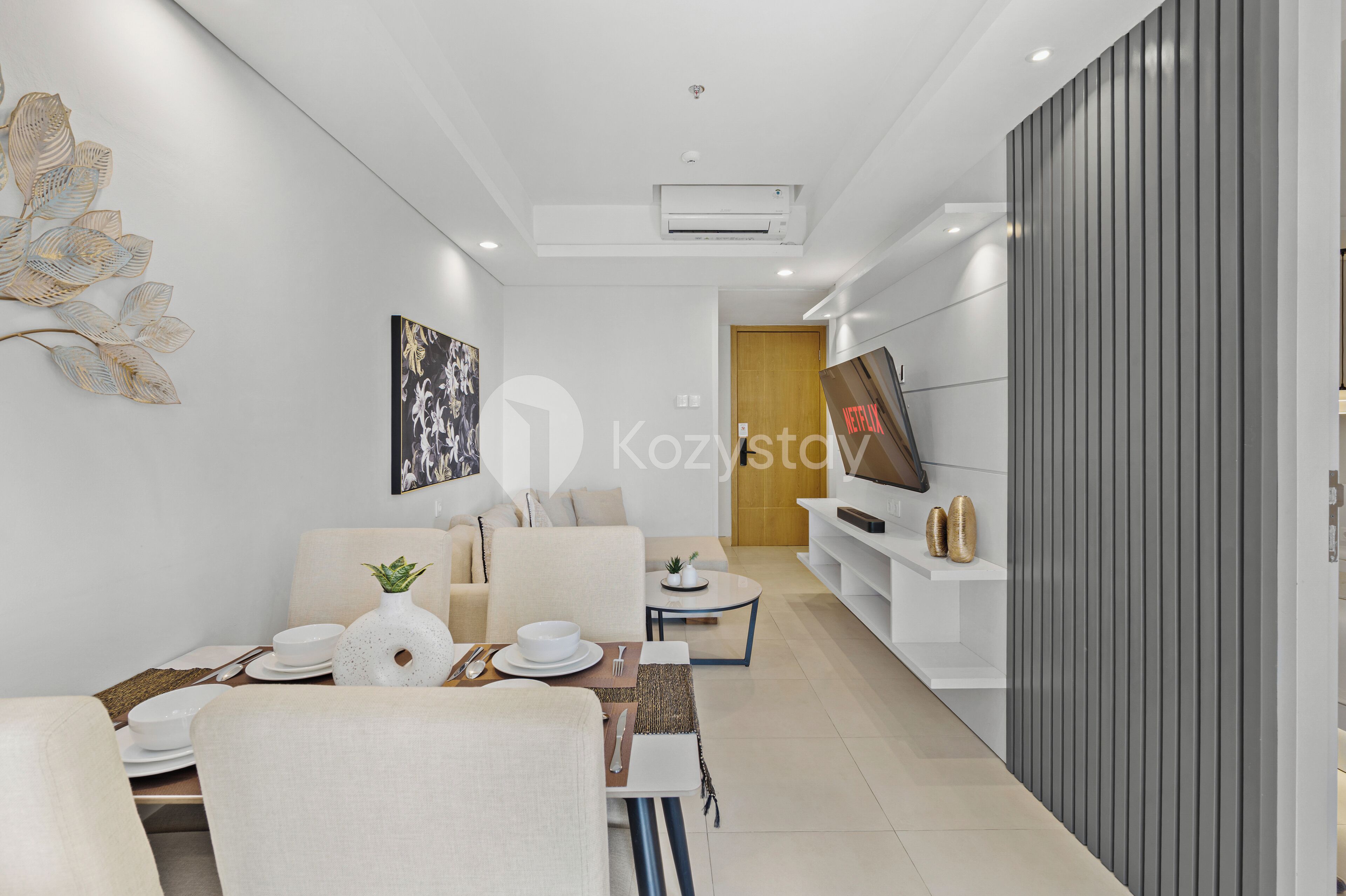 Una sala de estar moderna con un sofá, una mesa de centro y un comedor. Hay un televisor sobre un mueble bajo y una pieza decorativa en la pared.