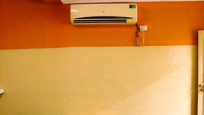 A room with an air conditioner mounted on an orange wall, a wooden bench, and a door on the right. - I-ROOMZ Surya Deluxe Lodge (Chikkaballapur)