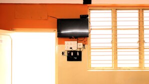 A room with a white refrigerator, a wall-mounted television, and a window with blinds. - I-ROOMZ Surya Deluxe Lodge (Chikkaballapur)