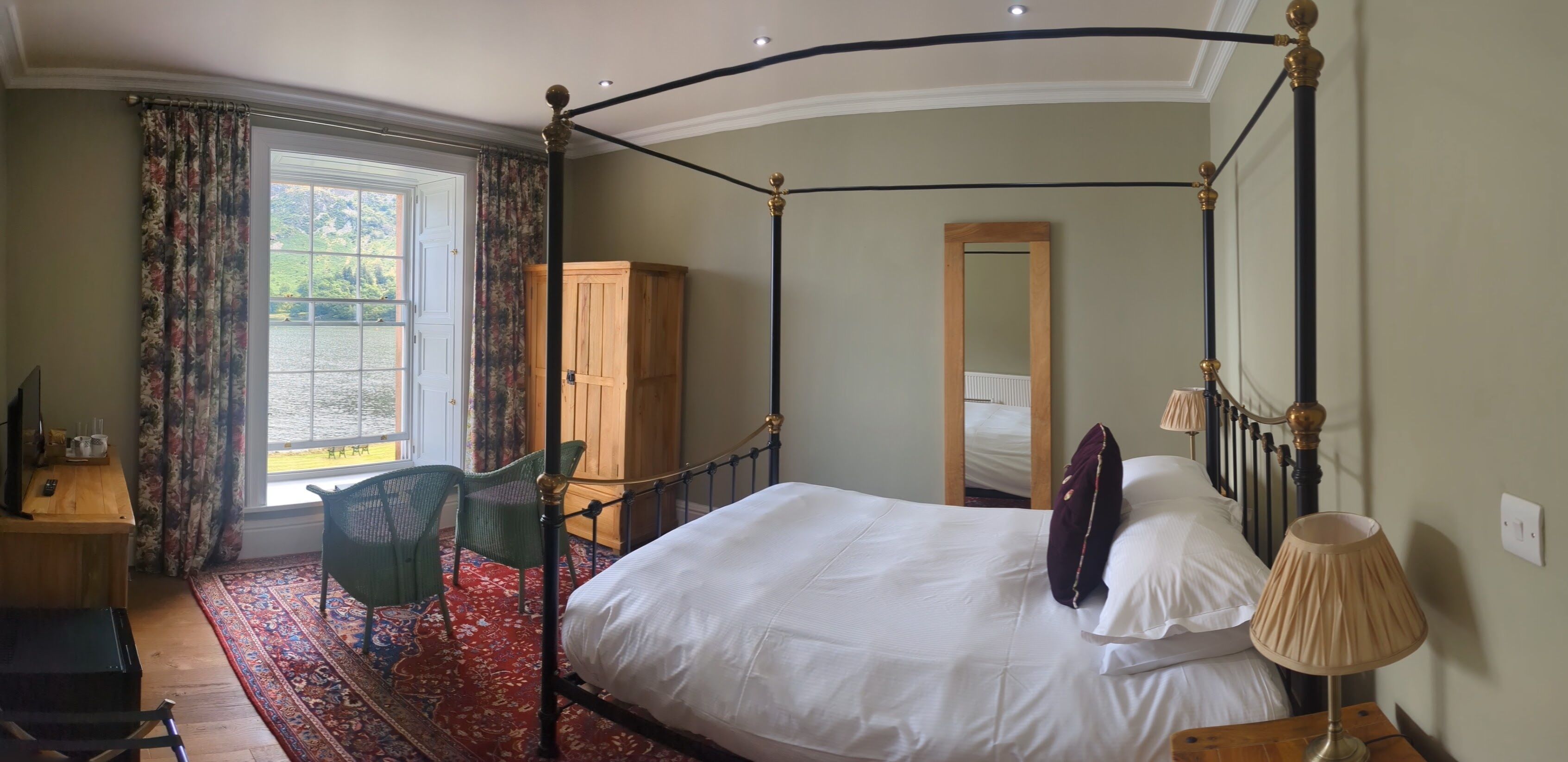 A four-poster bed with a wooden headboard, a mirror, and a lamp on a bedside table. A chair and a desk with a computer are visible in the corner.