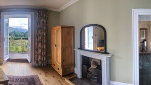 A bedroom with a four-poster bed, a wooden chest of drawers, a fireplace, and a view of a lake.
