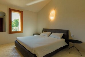 A bedroom with a bed, a small table, a nightstand, a window, and a wall-mounted light fixture.