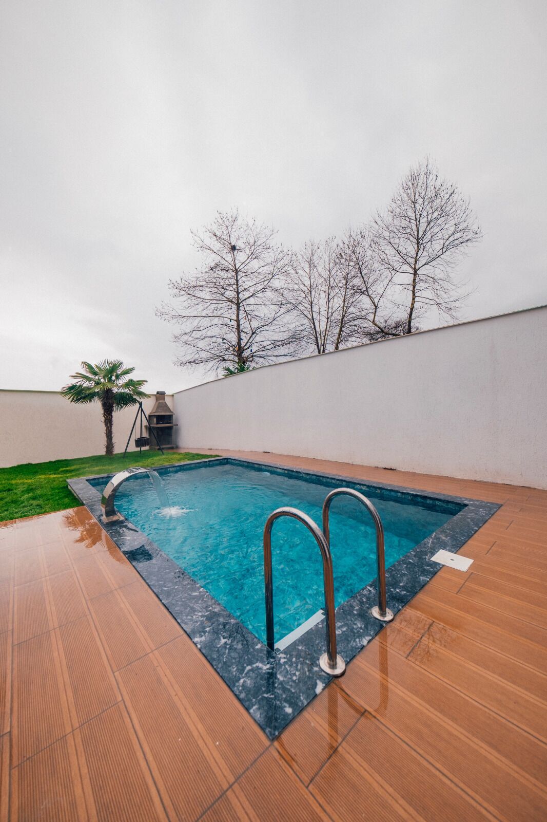 A rectangular swimming pool with a marble border, surrounded by a wooden deck and a low wall.