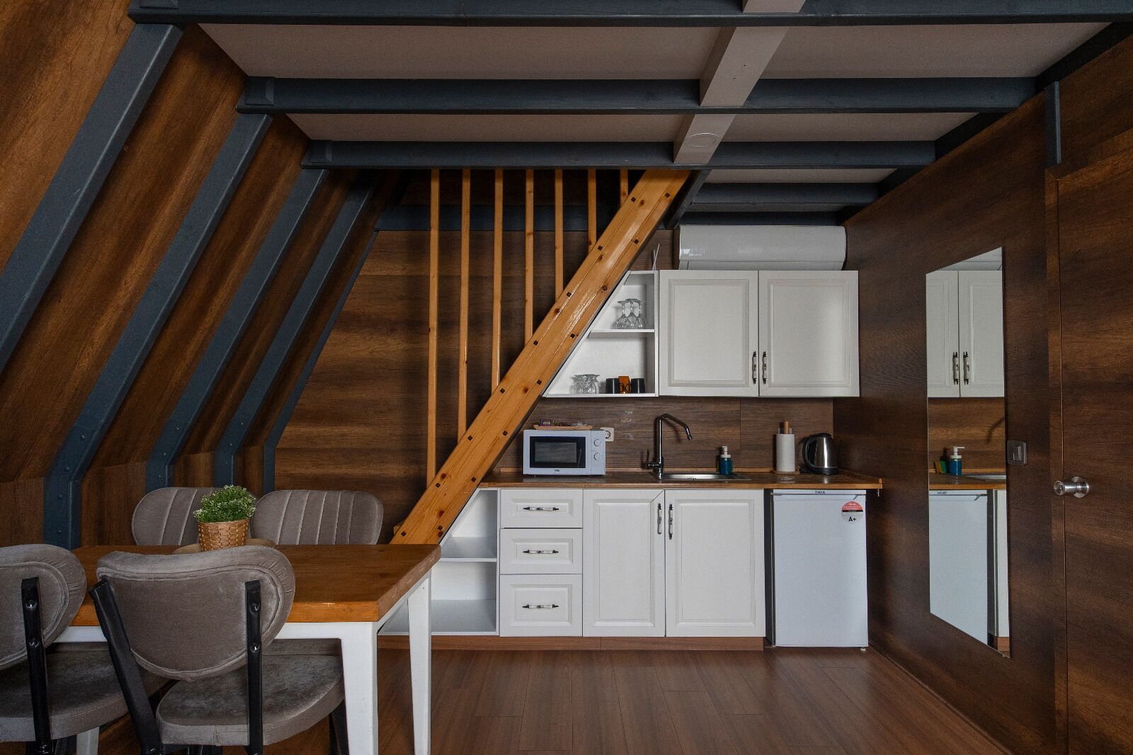 A compact kitchen with white cabinets, a wooden staircase, and a dining area with two chairs.