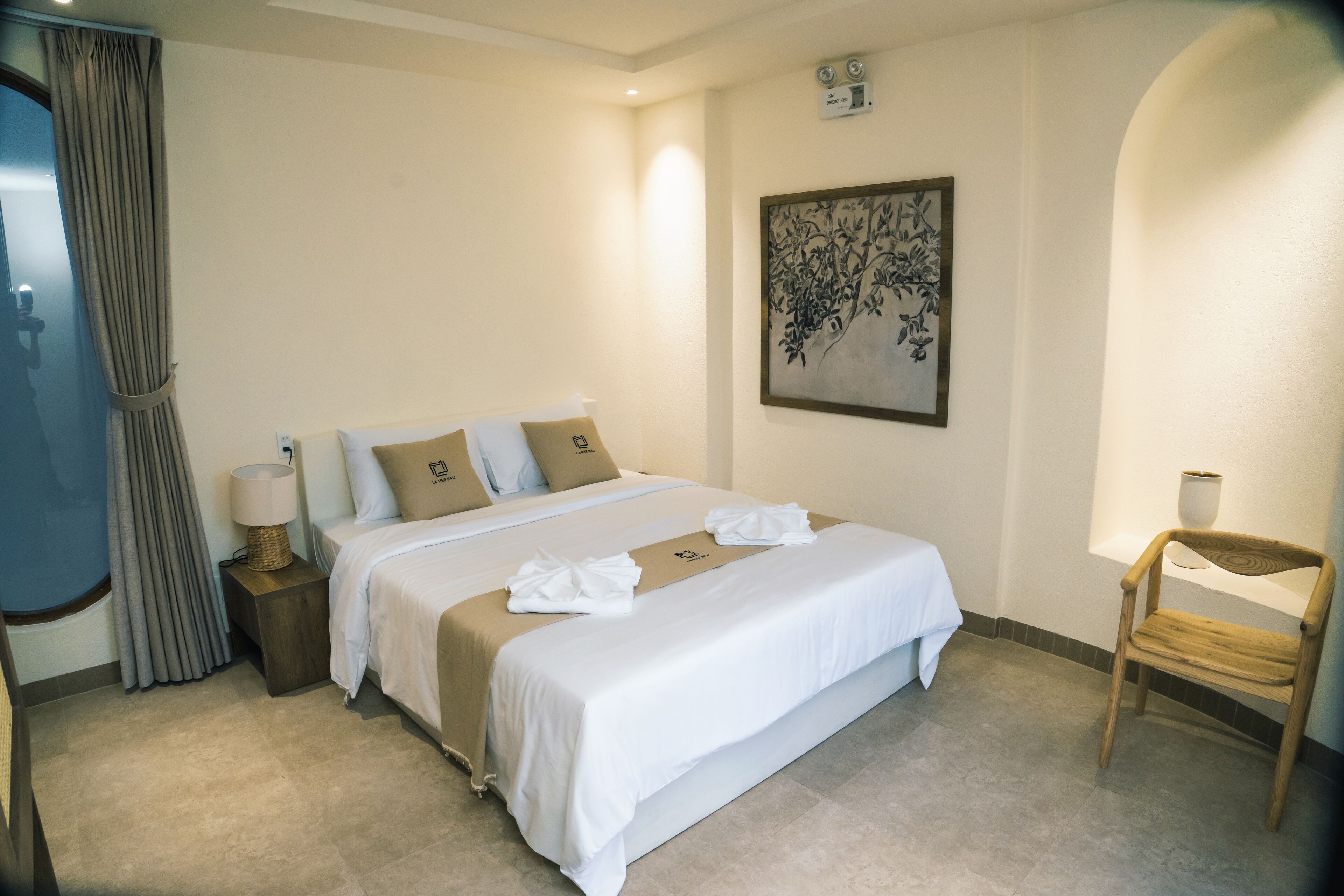 A hotel room with a bed, a nightstand with a lamp, a wooden chair, a painting on the wall, and a window with curtains.