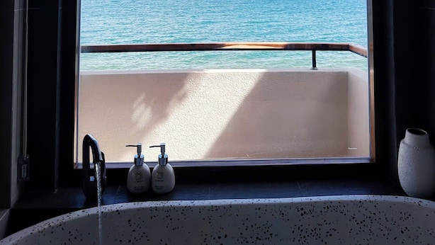 A bathroom with a large arched window overlooking the sea, a bathtub, and two bottles of shampoo. - La Mer Bali Mui Ne (Mui Ne)