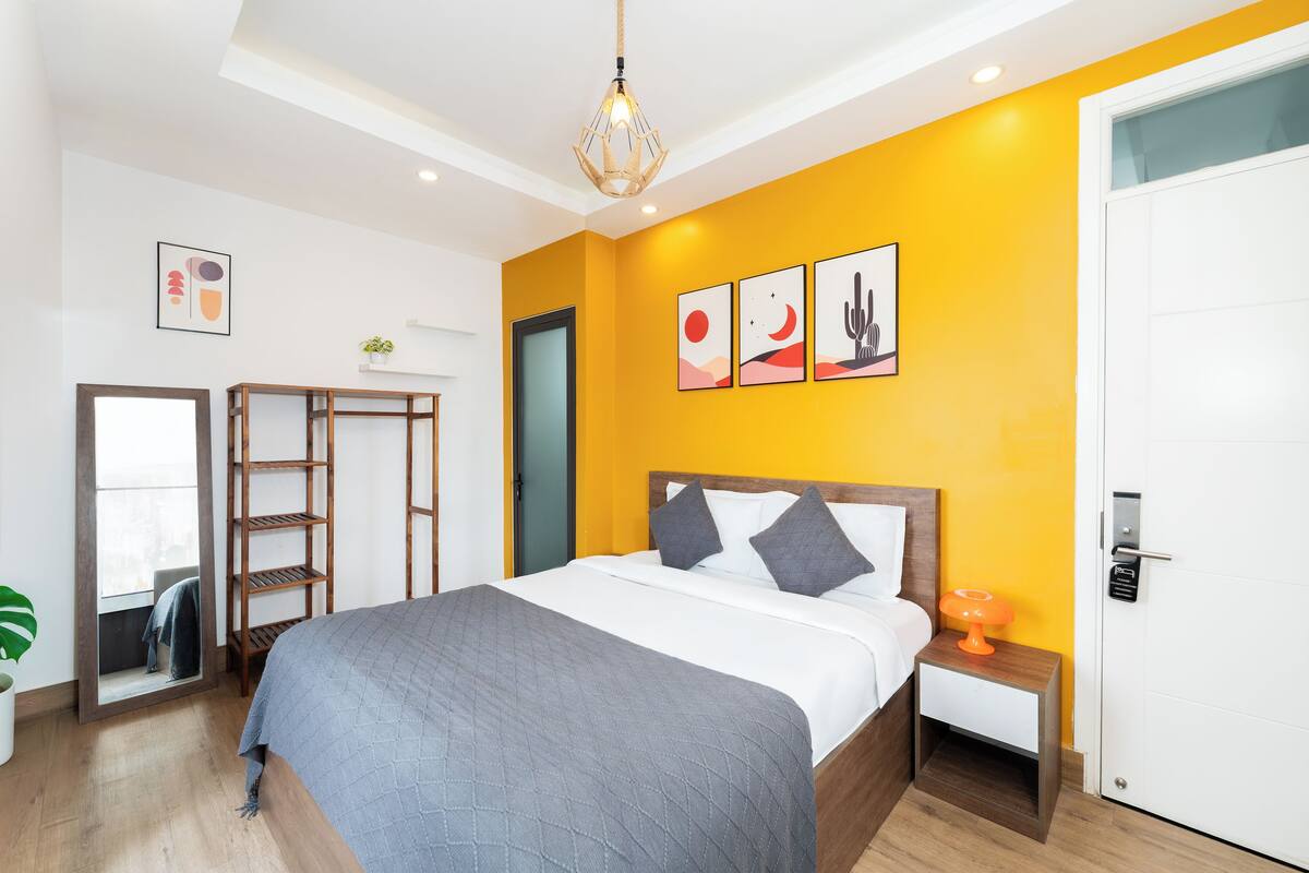 A modern hotel room with a large bed, a wooden headboard, a small bedside table with a lamp, and a bright yellow accent wall.