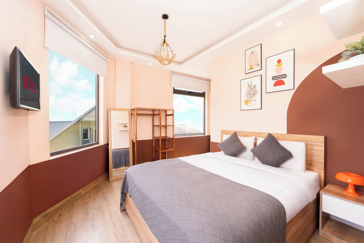 A modern hotel room with a large bed, a TV mounted on the wall, a window with a view, and a bedside table with a lamp.
