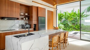A modern kitchen with a marble countertop, wooden cabinets, and a large island with a sink. There is a dining area with wicker chairs and a view of a lush garden through large glass doors. - De Dominique Villas Canggu (Canggu)