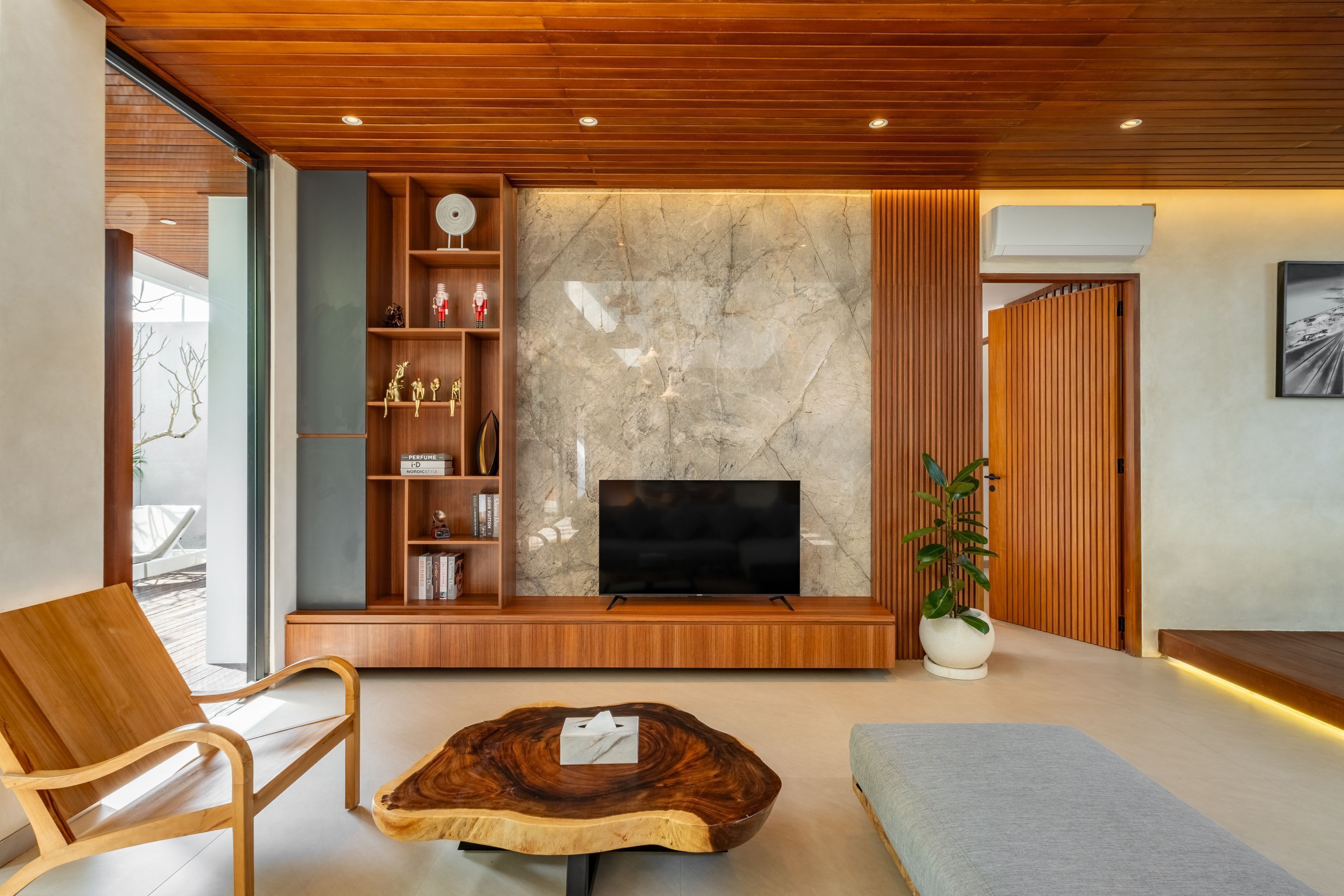 A modern living room with a wooden ceiling, a marble wall, a flat-screen TV, a wooden bookshelf, a plant, and a unique coffee table.