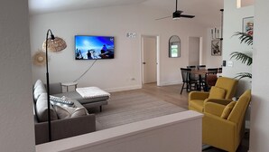 Ein modernes Wohnzimmer mit einer Couch, einem gelben Sessel, einem Essbereich mit Tisch und Stühlen, einem Deckenventilator und einem an der Wand montierten Fernseher.