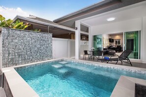 En modern utomhuspool med en mur av sten, solstolar och en matplats med bord och stolar.