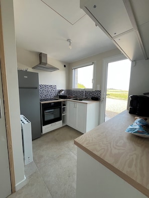 A compact kitchen with a built-in oven, a refrigerator, and a microwave. There is a window providing natural light and a view of the outdoors.