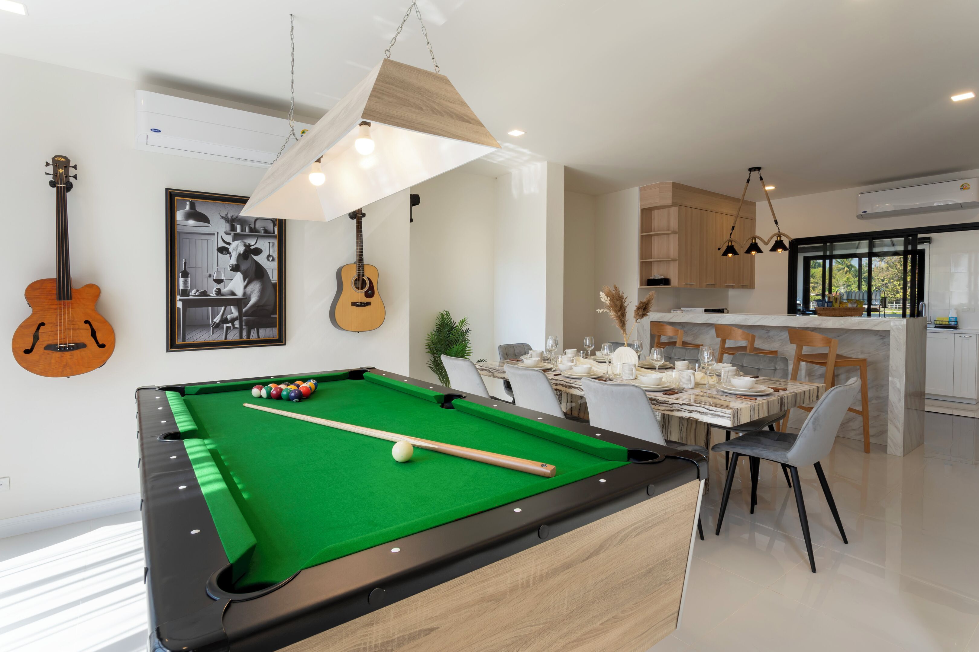 Un salon moderne avec un billard, un coin repas avec une table pour quatre personnes et une cuisine dotée de meubles en bois et d’un luminaire suspendu.