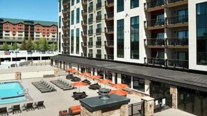 A rooftop pool area with lounge chairs and umbrellas, a stone fireplace, and a view of adjacent buildings.
