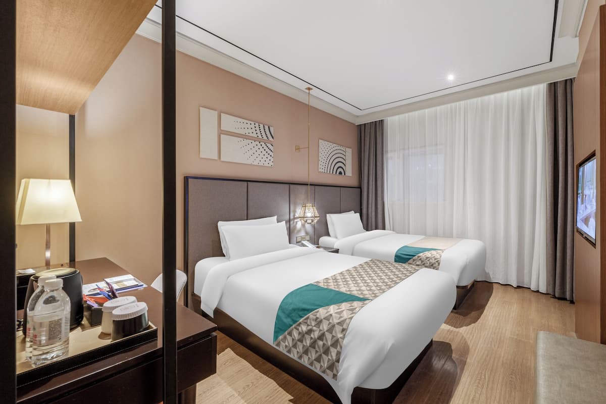 A hotel room with two beds, a desk with a lamp, and a large window with curtains.